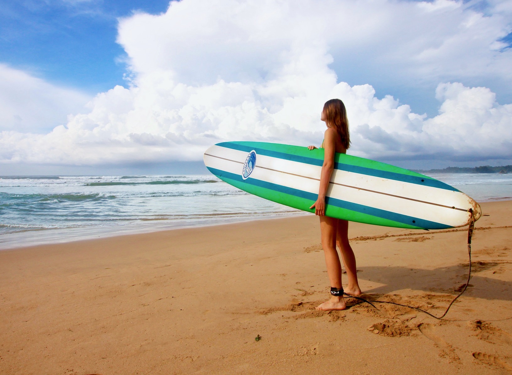 Una chica con una tabla de surf mirando al mar buscando olas antes de meterse en el agua.